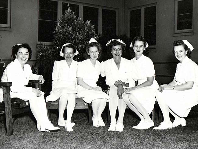6 smiling nurses seated on the lawn in front of the hospital