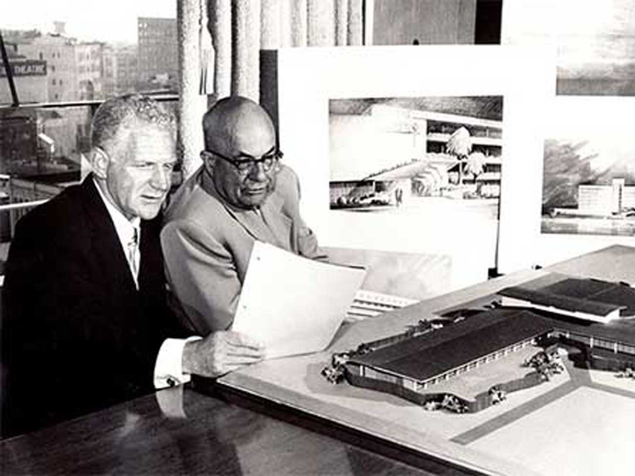 2 men wearing business suits looking over a miniature model of a hospital 